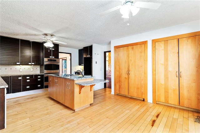 kitchen with stainless steel double oven, a kitchen island with sink, light wood-style flooring, and a kitchen bar