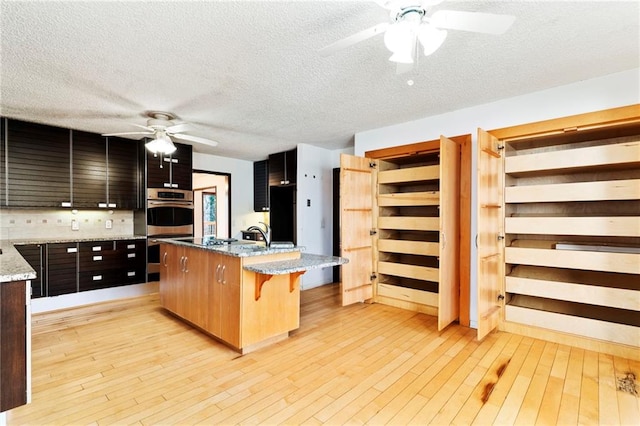 kitchen with a ceiling fan, a kitchen island with sink, light wood-style flooring, and a kitchen bar