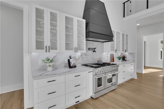 kitchen with backsplash, premium range hood, double oven range, white cabinetry, and light stone countertops