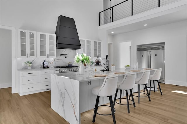 kitchen featuring premium range hood, white cabinetry, an island with sink, appliances with stainless steel finishes, and a kitchen breakfast bar