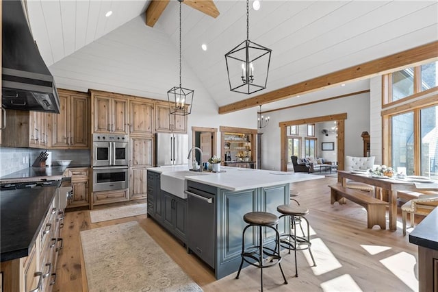 kitchen with an island with sink, stainless steel appliances, beamed ceiling, wall chimney exhaust hood, and light wood-type flooring