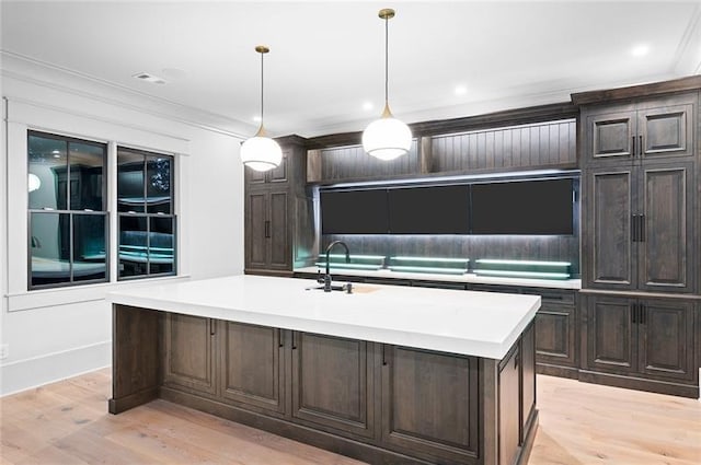 kitchen featuring light hardwood / wood-style flooring, crown molding, an island with sink, decorative light fixtures, and dark brown cabinets