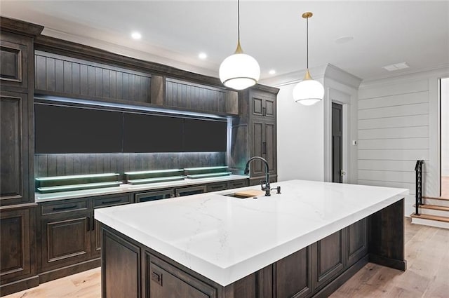 kitchen featuring sink, hanging light fixtures, wood walls, an island with sink, and light wood-type flooring