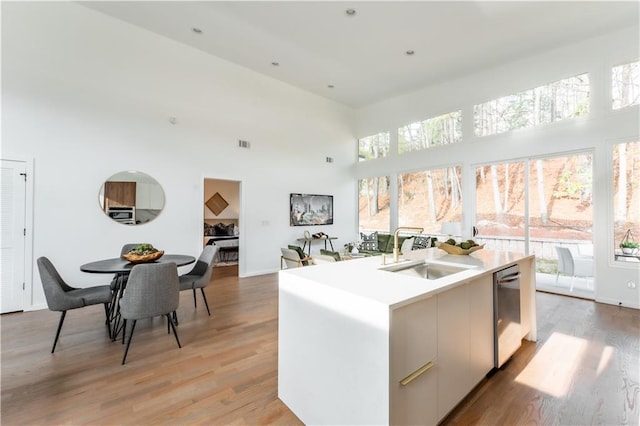 kitchen with a towering ceiling, stainless steel dishwasher, sink, light wood-type flooring, and a center island with sink