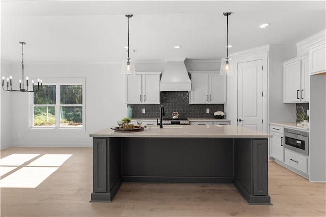 kitchen featuring custom exhaust hood, stainless steel microwave, white cabinetry, and a kitchen island with sink