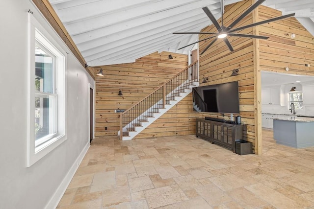 unfurnished living room featuring wooden walls, stone tile flooring, a ceiling fan, high vaulted ceiling, and a sink