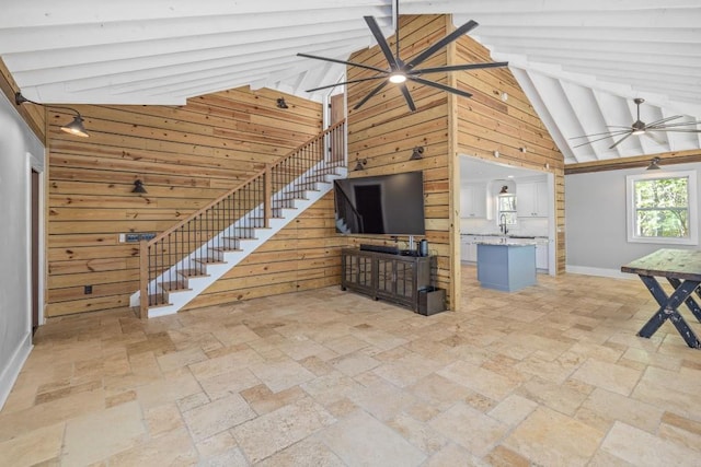 unfurnished living room with ceiling fan, stone tile flooring, high vaulted ceiling, wood walls, and baseboards