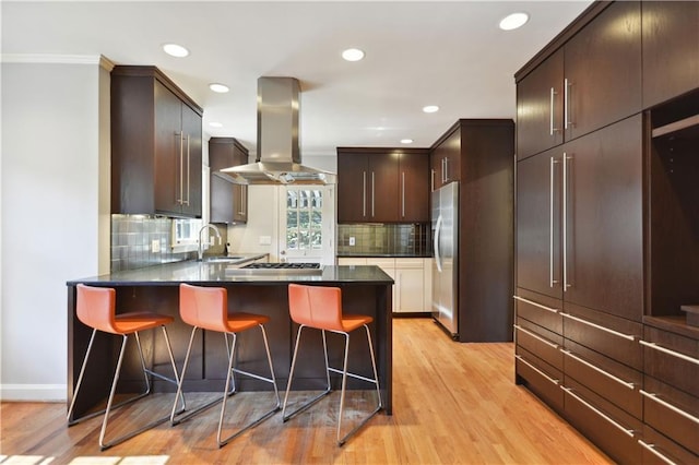 kitchen with dark countertops, a breakfast bar area, a peninsula, island exhaust hood, and light wood-style floors