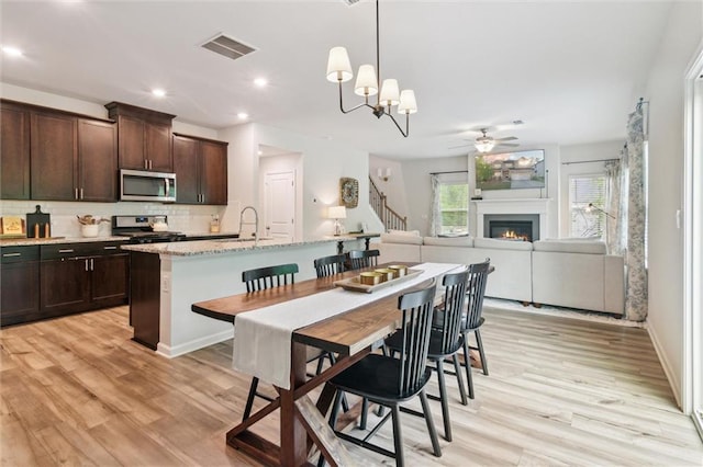 dining room with a warm lit fireplace, light wood finished floors, ceiling fan with notable chandelier, stairs, and visible vents