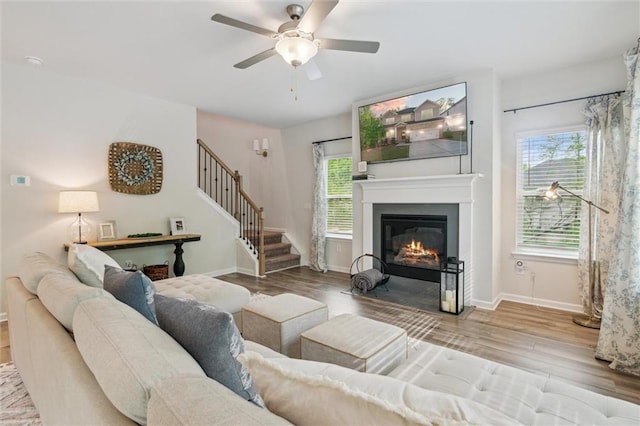 living area with a ceiling fan, wood finished floors, baseboards, a glass covered fireplace, and stairs
