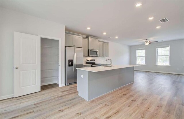 kitchen featuring light wood-type flooring, ceiling fan, an island with sink, appliances with stainless steel finishes, and gray cabinetry