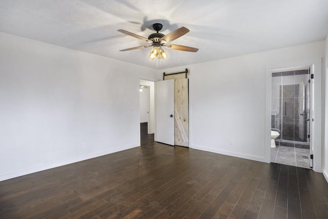 unfurnished bedroom featuring a ceiling fan, a barn door, baseboards, ensuite bath, and dark wood-style floors