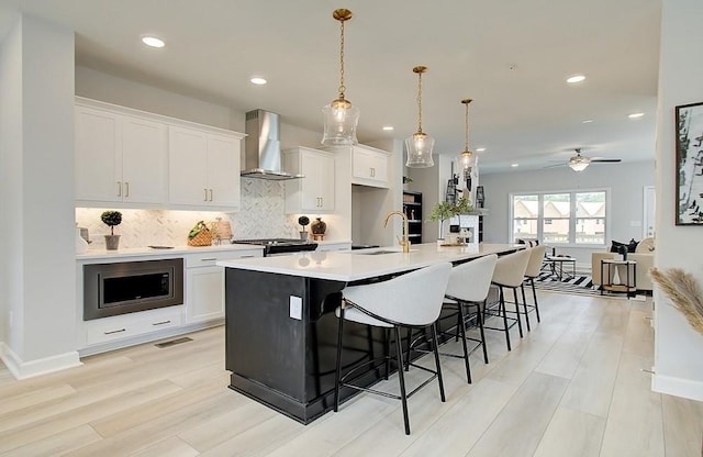 kitchen with white cabinetry, wall chimney range hood, an island with sink, hanging light fixtures, and stainless steel microwave