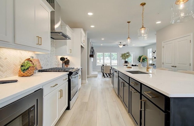 kitchen featuring ceiling fan, wall chimney exhaust hood, white cabinets, an island with sink, and stainless steel appliances