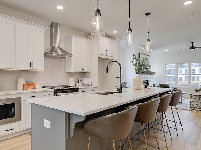 kitchen with a kitchen island with sink, wall chimney range hood, white cabinets, sink, and stainless steel range with gas stovetop