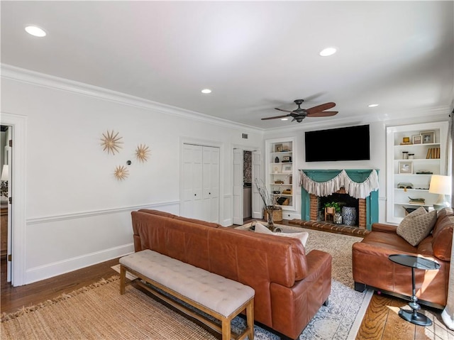 living area with a ceiling fan, a fireplace, built in shelves, wood finished floors, and ornamental molding