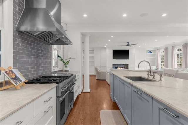 kitchen with a sink, double oven range, a warm lit fireplace, wall chimney range hood, and ceiling fan