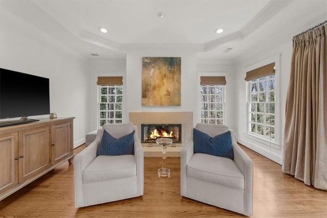 living area featuring light wood-type flooring, a warm lit fireplace, a raised ceiling, and crown molding