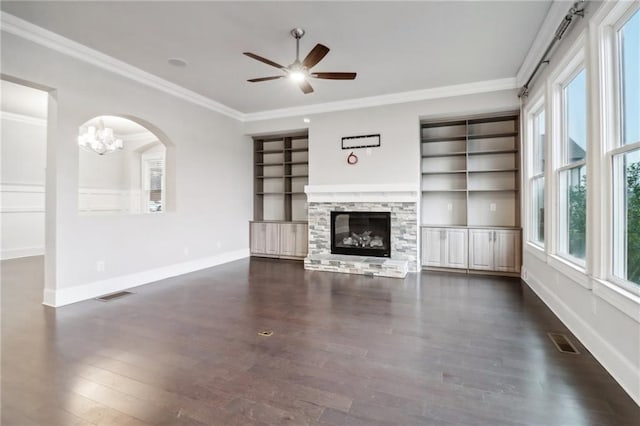 unfurnished living room with ceiling fan with notable chandelier, crown molding, dark wood-type flooring, and a stone fireplace