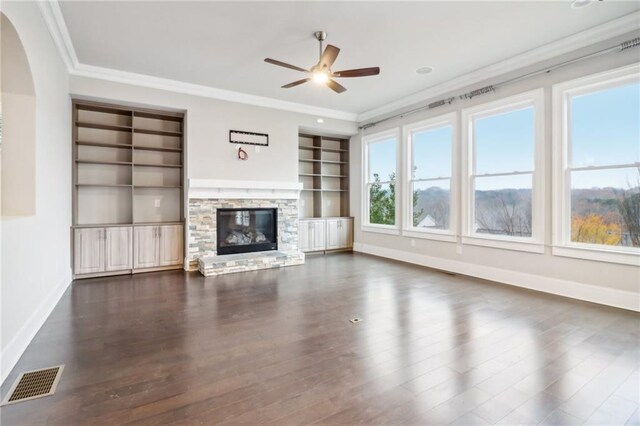 unfurnished living room featuring ceiling fan, a stone fireplace, built in features, dark hardwood / wood-style floors, and ornamental molding