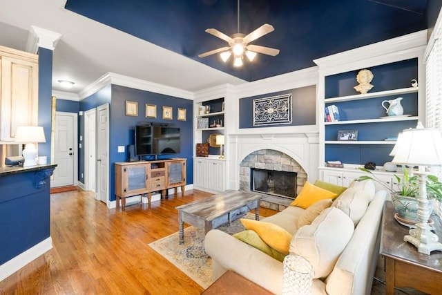 living area featuring ceiling fan, light wood-style flooring, ornamental molding, a stone fireplace, and baseboards