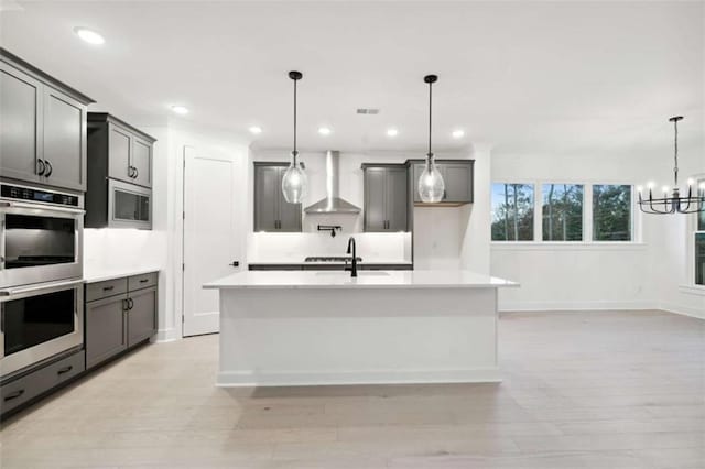 kitchen with an inviting chandelier, wall chimney exhaust hood, appliances with stainless steel finishes, light countertops, and a sink