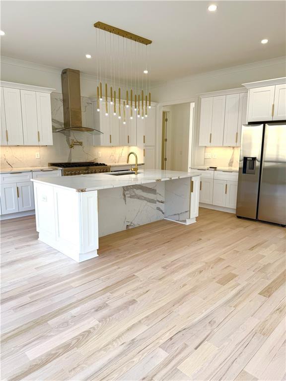 kitchen featuring stainless steel refrigerator with ice dispenser, wall chimney exhaust hood, a kitchen island with sink, and hanging light fixtures