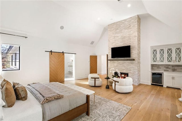 bedroom with a fireplace, a dry bar, light wood-style flooring, a barn door, and high vaulted ceiling