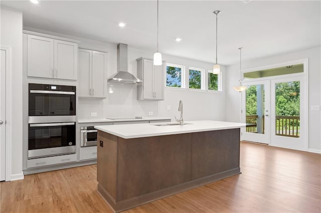 kitchen with light countertops, light wood-style floors, stainless steel double oven, a sink, and wall chimney exhaust hood