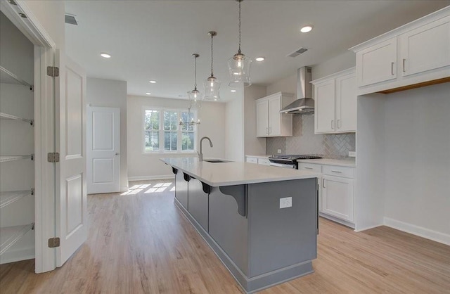 kitchen with wall chimney exhaust hood, sink, hanging light fixtures, an island with sink, and white cabinets