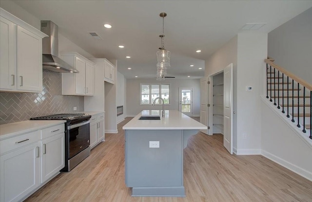 kitchen with sink, stainless steel gas range, white cabinetry, a center island with sink, and wall chimney exhaust hood