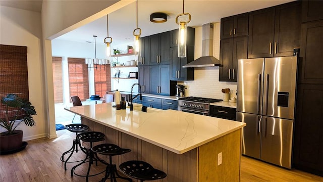 kitchen with an island with sink, light wood-style flooring, stainless steel appliances, a breakfast bar area, and wall chimney exhaust hood