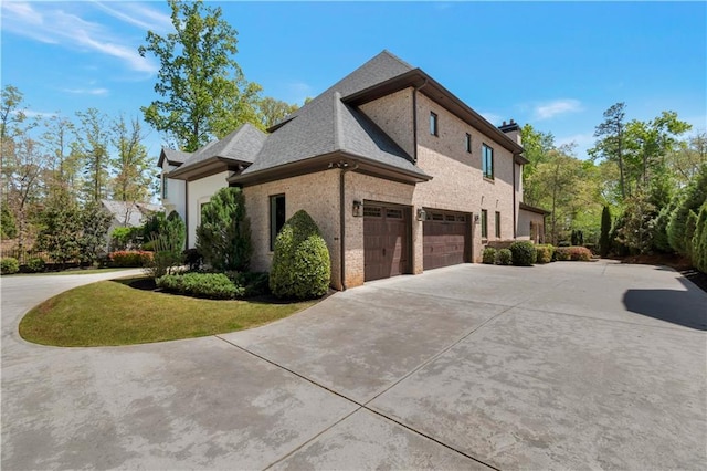 view of side of home with driveway, brick siding, an attached garage, and a chimney