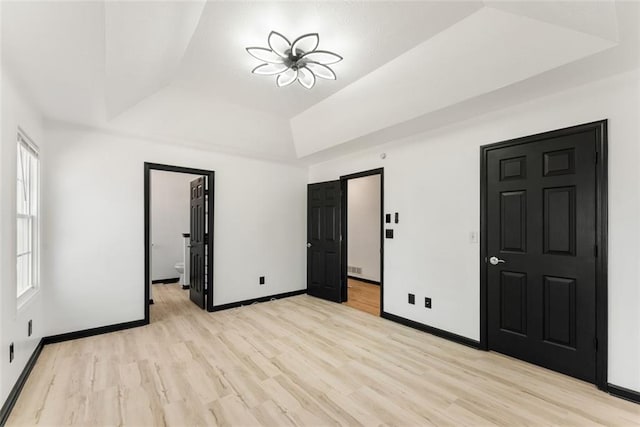 unfurnished bedroom featuring a tray ceiling, ensuite bath, light wood-style floors, and baseboards