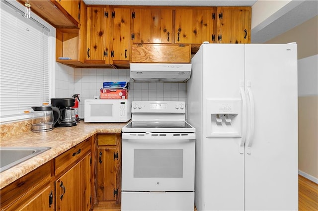 kitchen featuring under cabinet range hood, white appliances, light countertops, decorative backsplash, and brown cabinetry