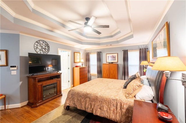 bedroom with a tray ceiling, multiple windows, ornamental molding, light wood-type flooring, and a glass covered fireplace
