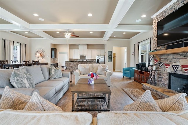 living room with coffered ceiling, ceiling fan, light hardwood / wood-style flooring, beamed ceiling, and a stone fireplace