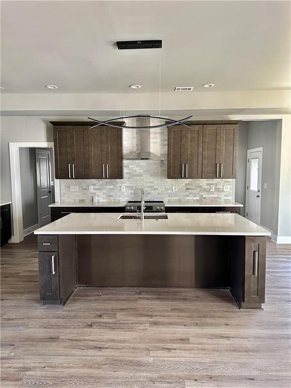 kitchen with dark brown cabinetry, sink, light hardwood / wood-style flooring, a center island with sink, and wall chimney range hood