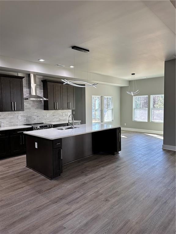 kitchen featuring pendant lighting, wall chimney range hood, sink, dark brown cabinets, and a center island with sink