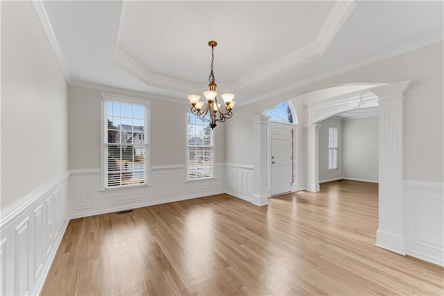 unfurnished dining area featuring light wood finished floors, a chandelier, arched walkways, a raised ceiling, and ornate columns