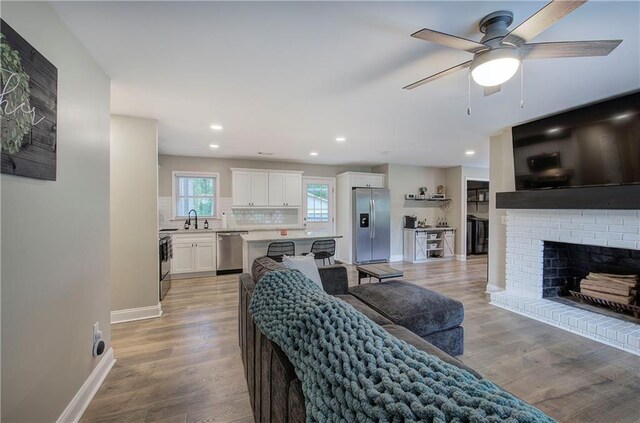 living area with ceiling fan, a brick fireplace, baseboards, light wood finished floors, and recessed lighting