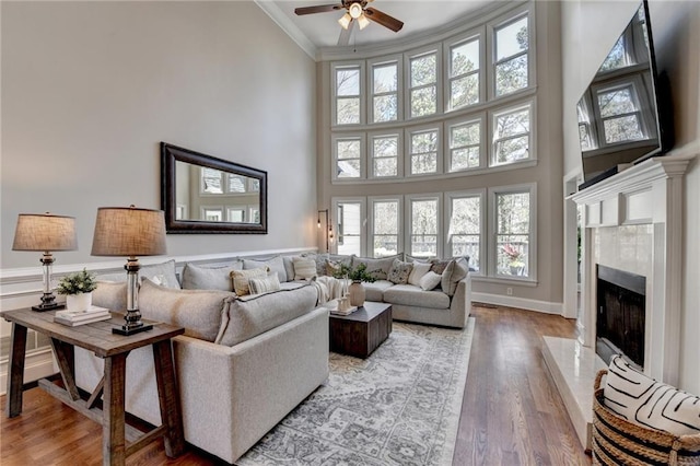 living room with ceiling fan, ornamental molding, a fireplace, a towering ceiling, and wood finished floors