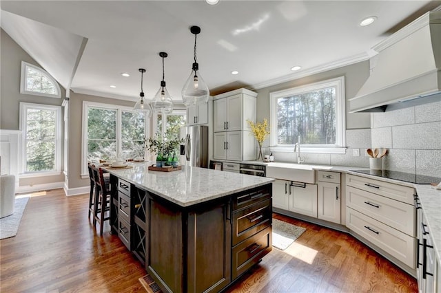 kitchen with custom range hood, stainless steel refrigerator with ice dispenser, wood finished floors, black electric cooktop, and a sink