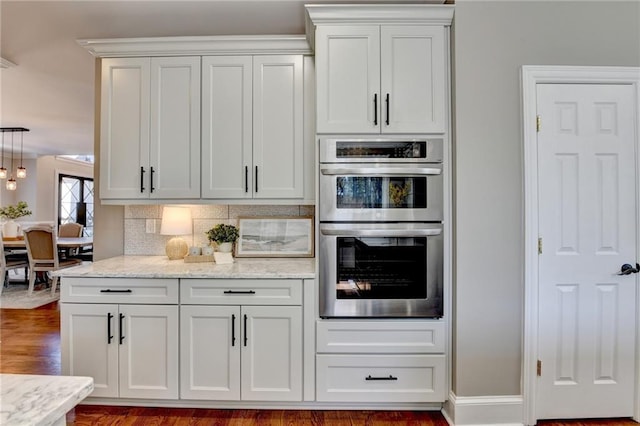 kitchen with light stone countertops, stainless steel double oven, dark wood-type flooring, white cabinetry, and tasteful backsplash