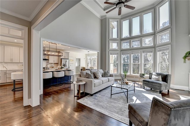 living area with dark wood-style flooring, a towering ceiling, ornamental molding, baseboards, and ceiling fan with notable chandelier