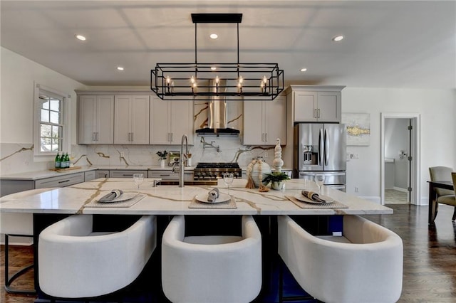 kitchen featuring light stone counters, a breakfast bar, stainless steel refrigerator with ice dispenser, a sink, and wall chimney range hood