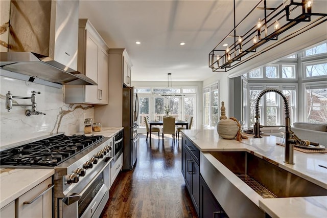 kitchen featuring stainless steel appliances, hanging light fixtures, a sink, wall chimney range hood, and light stone countertops