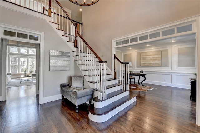 staircase featuring baseboards, wood finished floors, a high ceiling, a chandelier, and a decorative wall