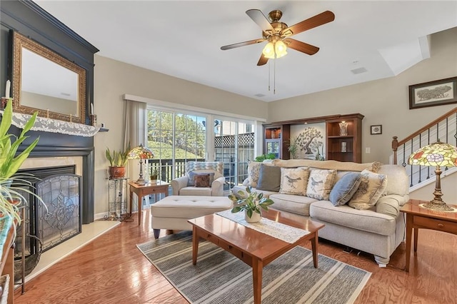 living area featuring baseboards, a fireplace with raised hearth, ceiling fan, wood finished floors, and stairs