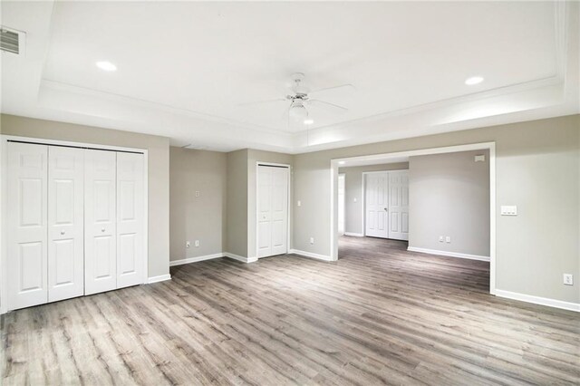 unfurnished bedroom with ceiling fan, a raised ceiling, wood-type flooring, and multiple closets